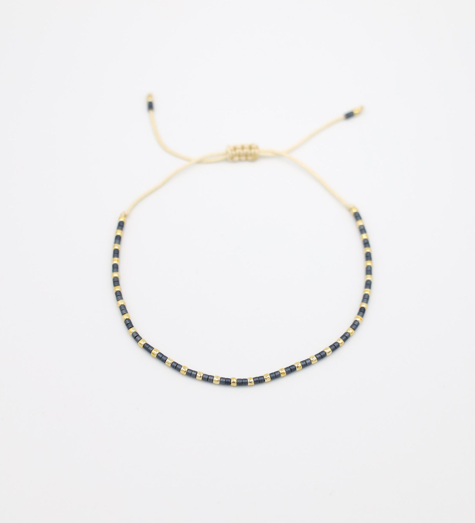 Flat-lay image of the Anya bracelet featuring alternating dark and gold beads, strung on a cream cord with an adjustable sliding closure.