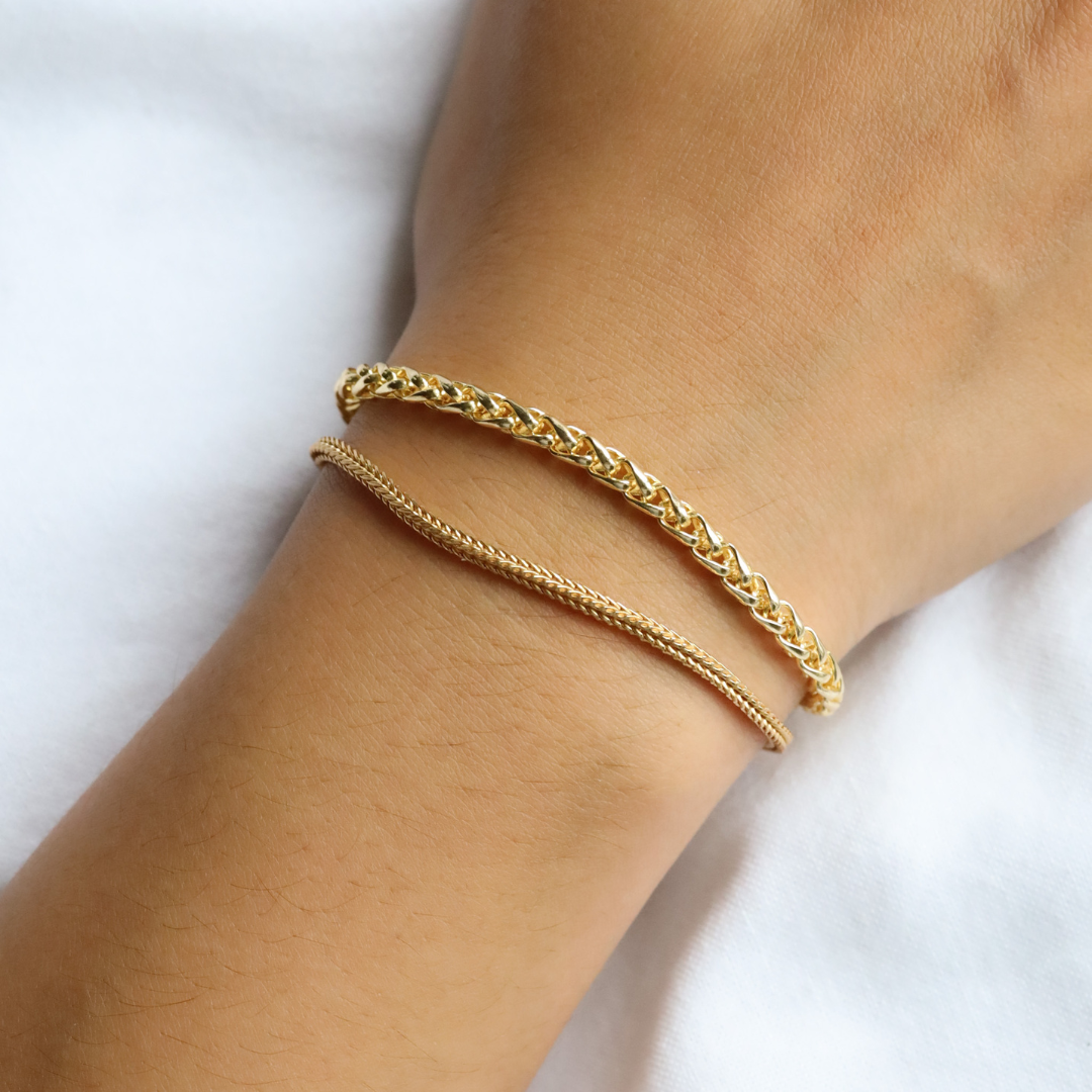 Two minimalist chain bracelets worn on wrist – one sleek wheat chain and one round braided chain, both made of 925 sterling silver with gold finish.
