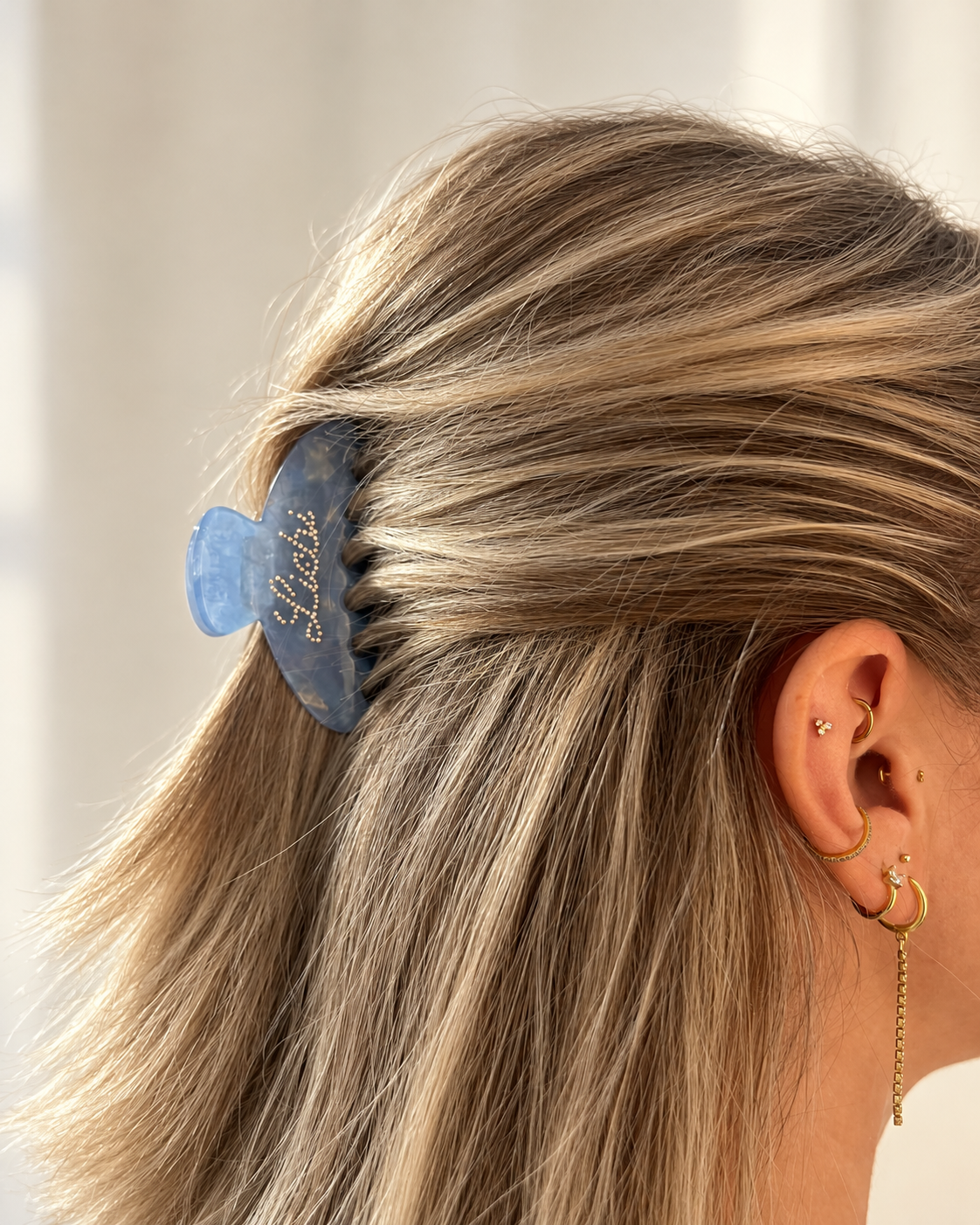 A close-up side view of a person's head wearing a translucent blue hair claw clip with gold script lettering, styled with blonde hair and gold hoop earrings.