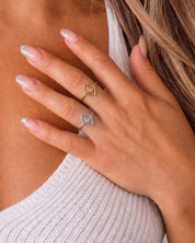 A close-up of a woman's hand resting on her chest, wearing two rings: a gold ring with a small pink stone and a silver ring with a small blue stone.