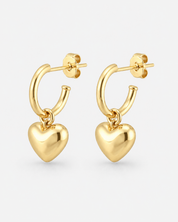 A pair of gold hoop earrings with dangling polished heart charms, shown on a light gray background.