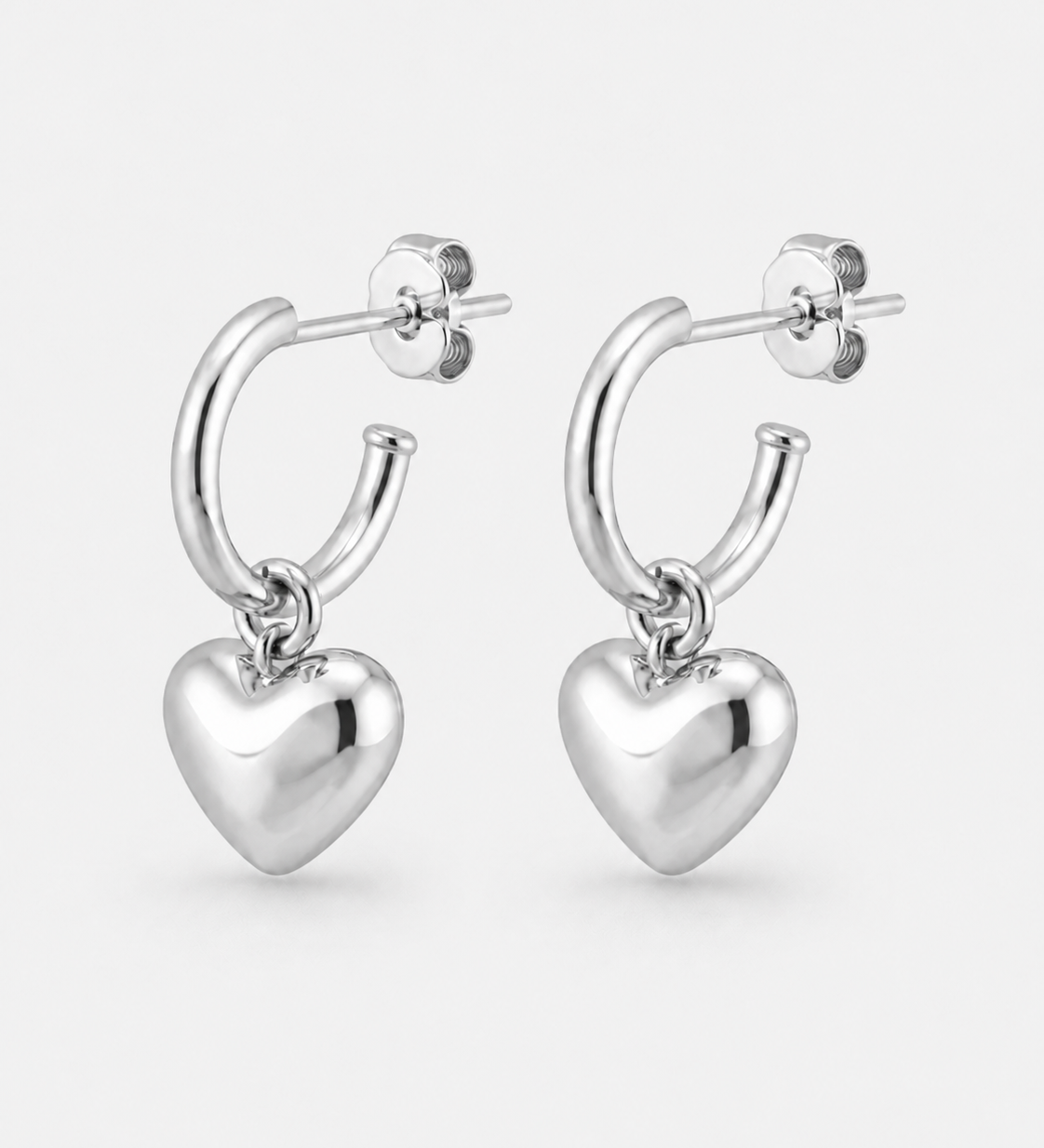 A pair of polished silver huggie hoop earrings with dangling heart charms, shown on a light gray background.