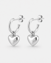 A pair of polished silver huggie hoop earrings with dangling heart charms, shown on a light gray background.